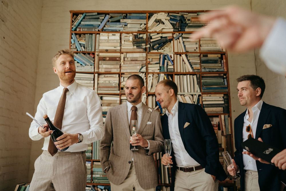 Four men in casual suits laughing together in front of a wall of stacked books.