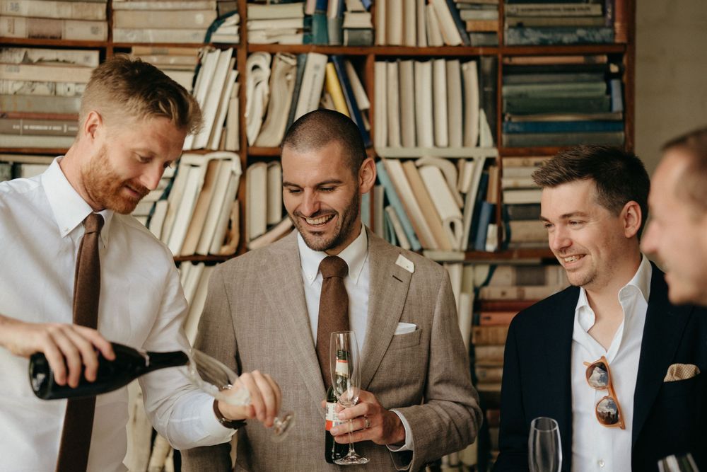 Groomsmen pour and share drinks while laughing in front of a bookshelf.