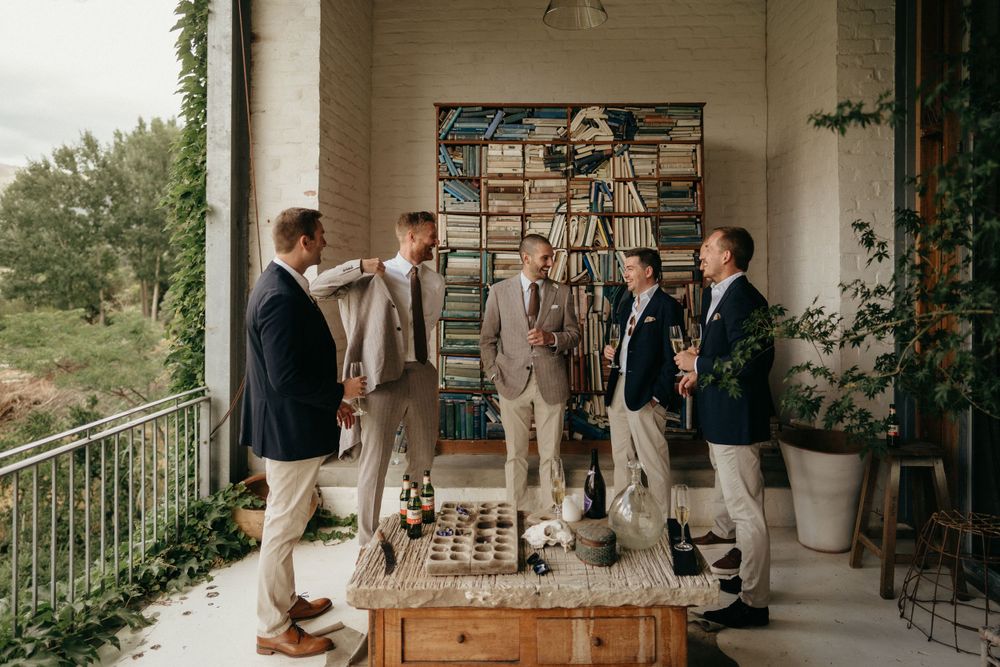 Groomsmen in casual suits gather on a porch with bookshelves and natural lighting.