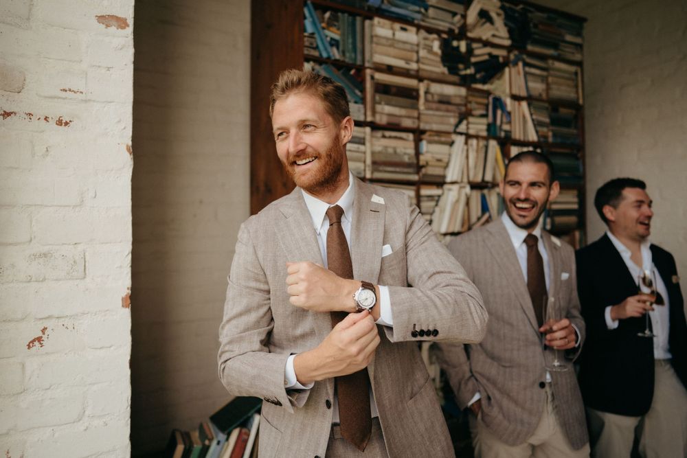 Wedding guests in suits share laughter and drinks during a casual reception moment.