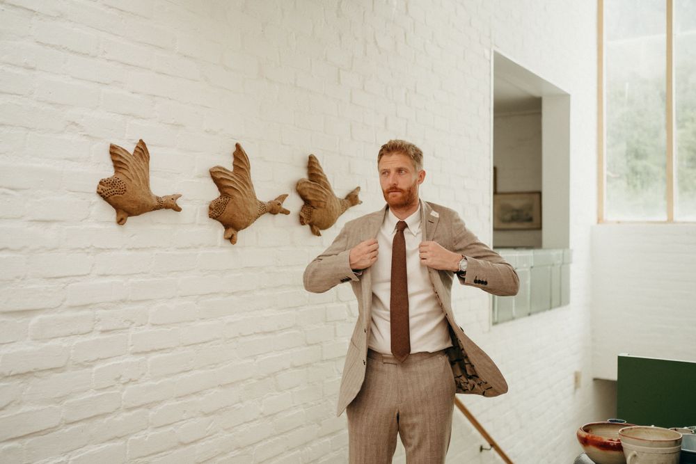 Man in beige suit poses playfully against white wall with decorative wooden duck decor.