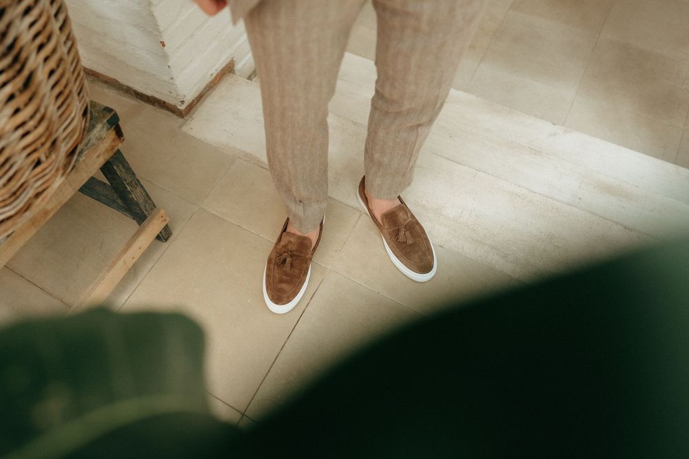 Brown leather slip-on shoes worn with light colored pants on a bright indoor floor.