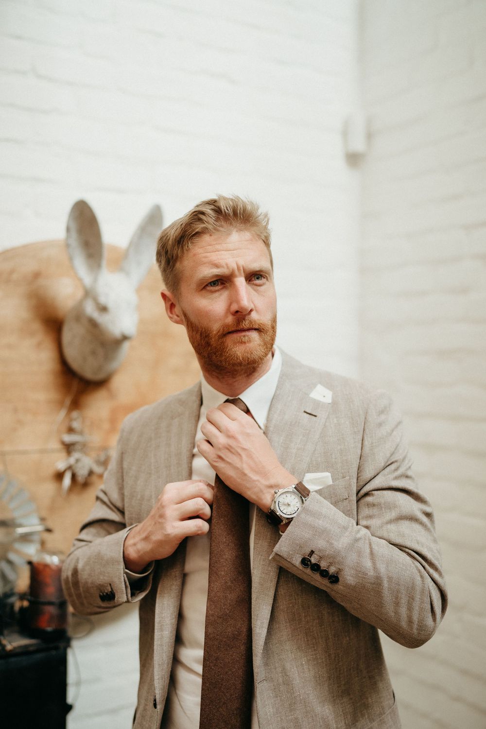 Person in light gray suit adjusts tie while standing against white wall with decorative rabbit head.