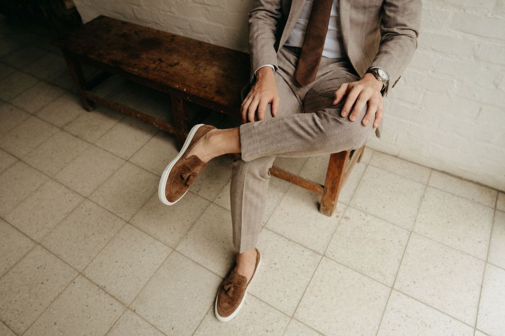 A person wearing tan pants and brown loafers sits on a wooden bench against a white wall.