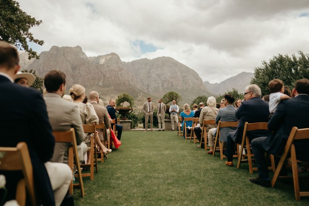 Mountain backdrop frames outdoor wedding ceremony with guests seated on wooden chairs on a grassy lawn.