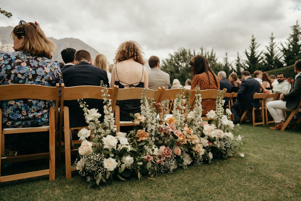 Wedding guests seated in wooden chairs with floral arrangements decorating the aisle at an outdoor ceremony.