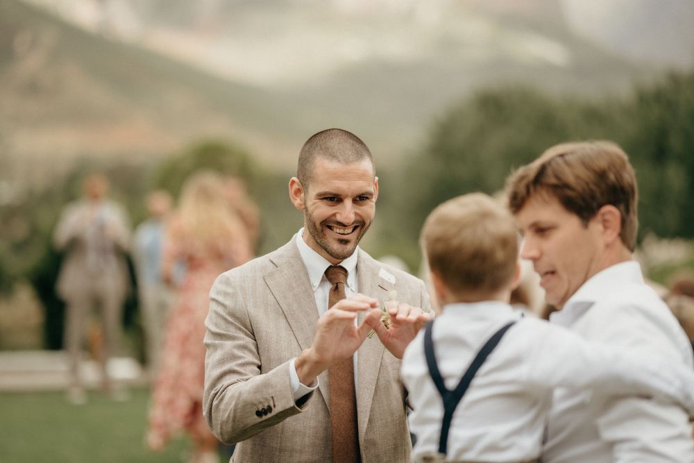 Wedding guests in light tan suit and suspenders sharing a light moment at an outdoor mountain venue.