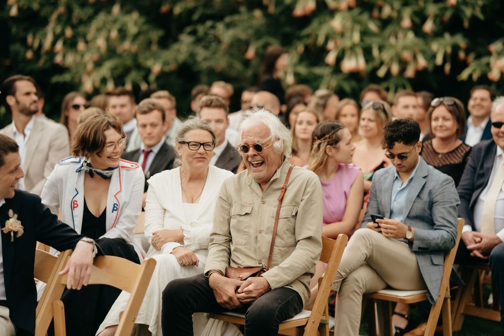 Attendees seated at an outdoor wedding ceremony in white chairs with leafy green foliage in the background.