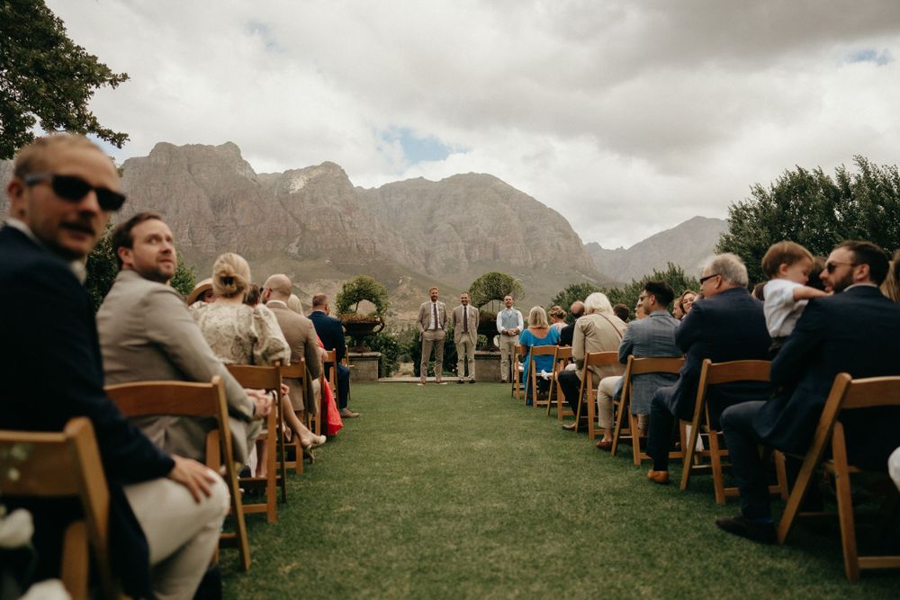 Outdoor wedding ceremony setup with wooden chairs on grass facing dramatic mountain landscape under cloudy skies.