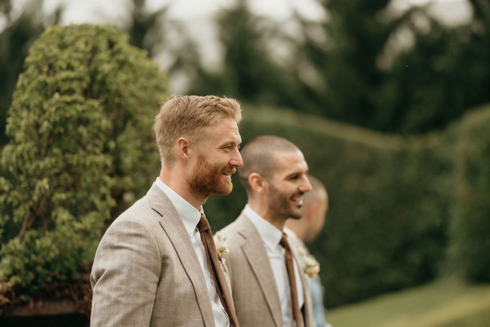 Two groomsmen in light gray suits smile during an outdoor wedding ceremony.