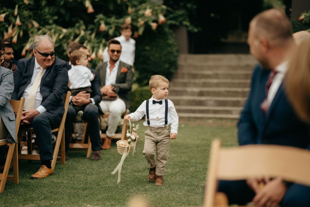 A small child wearing suspenders walks down the aisle at an outdoor wedding ceremony.