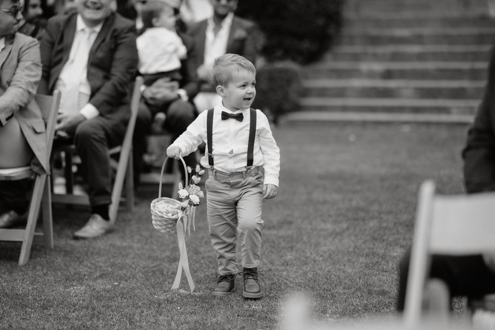 A young ring bearer in suspenders walks down the ceremony aisle carrying a basket.