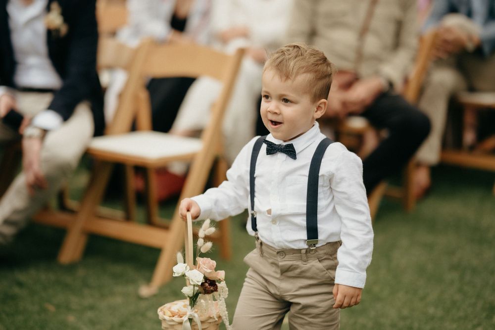 A young ring bearer in suspenders and bow tie carries a flower basket at an outdoor wedding ceremony.