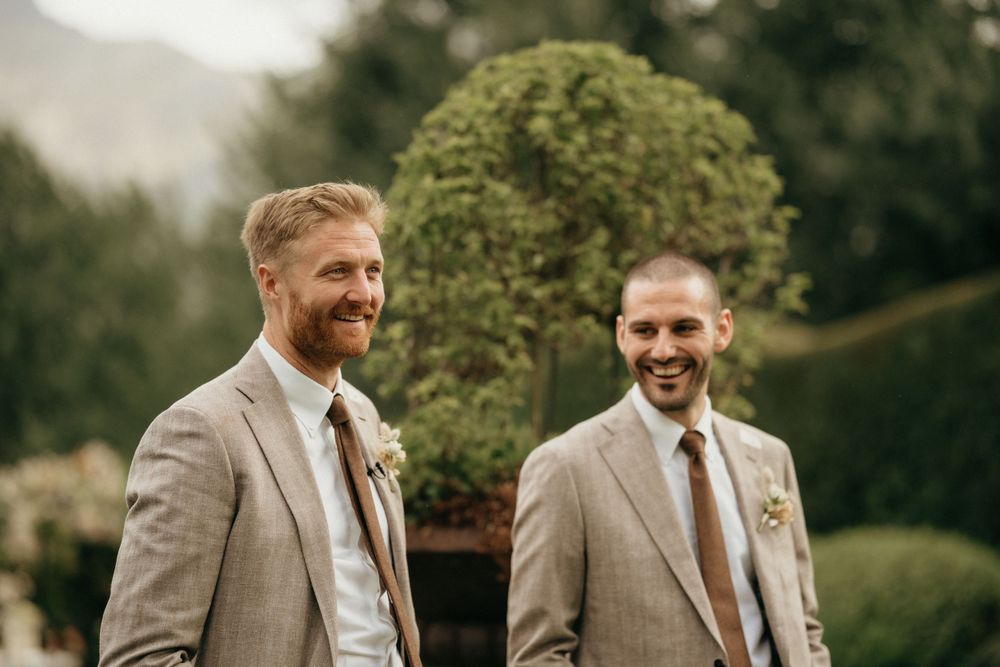 Two groomsmen in light gray suits and white boutonnieres share a joyful moment at an outdoor wedding.