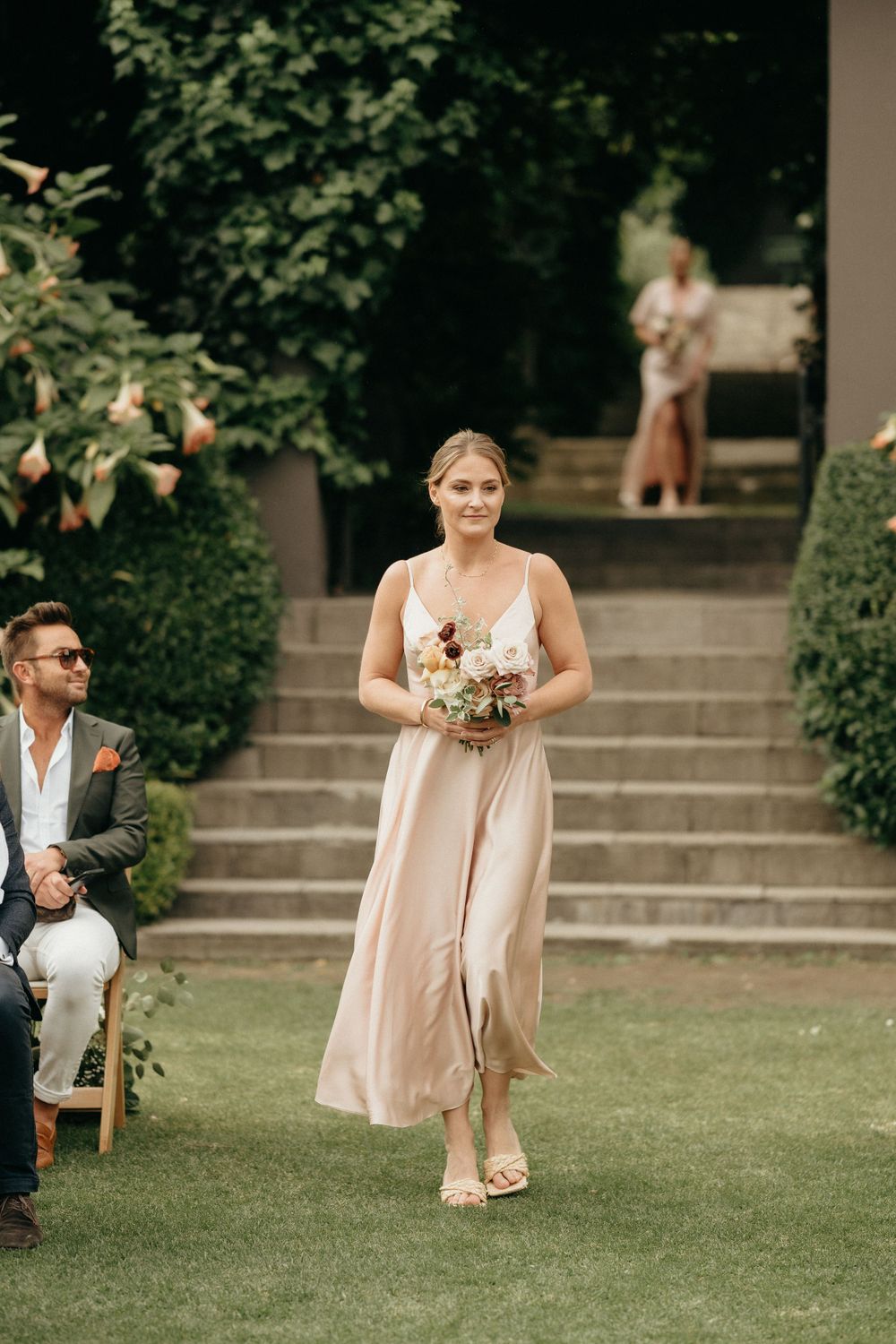 Wedding attendee in a blush gown walks down outdoor stone steps carrying a floral bouquet.