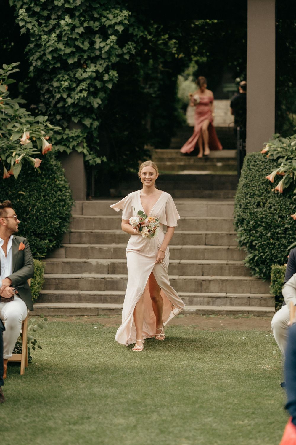 A bridesmaid in flowing champagne dress descends garden stairs during an outdoor ceremony.