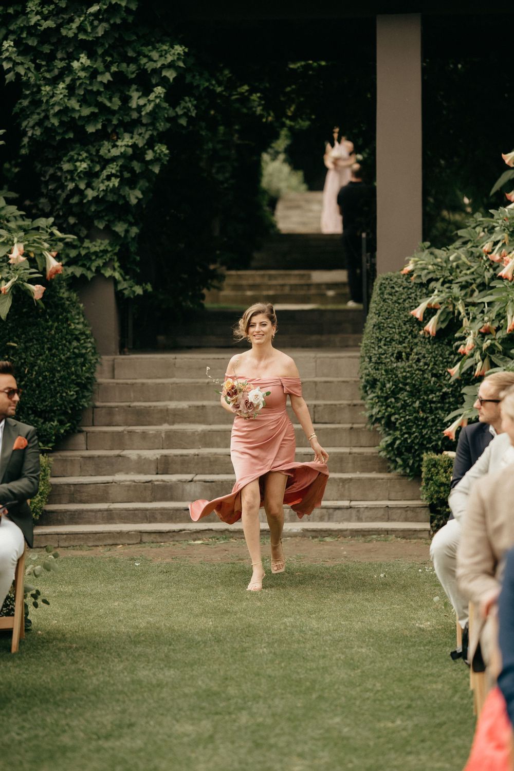 A bridesmaid in a pink dress descends stone stairs at an outdoor wedding ceremony.