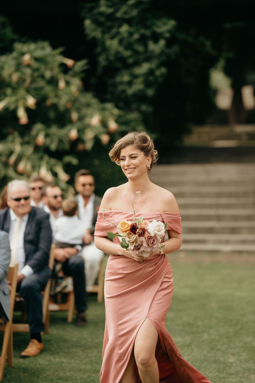 Wedding party member in flowing peach gown carries bouquet down grassy aisle at outdoor ceremony.