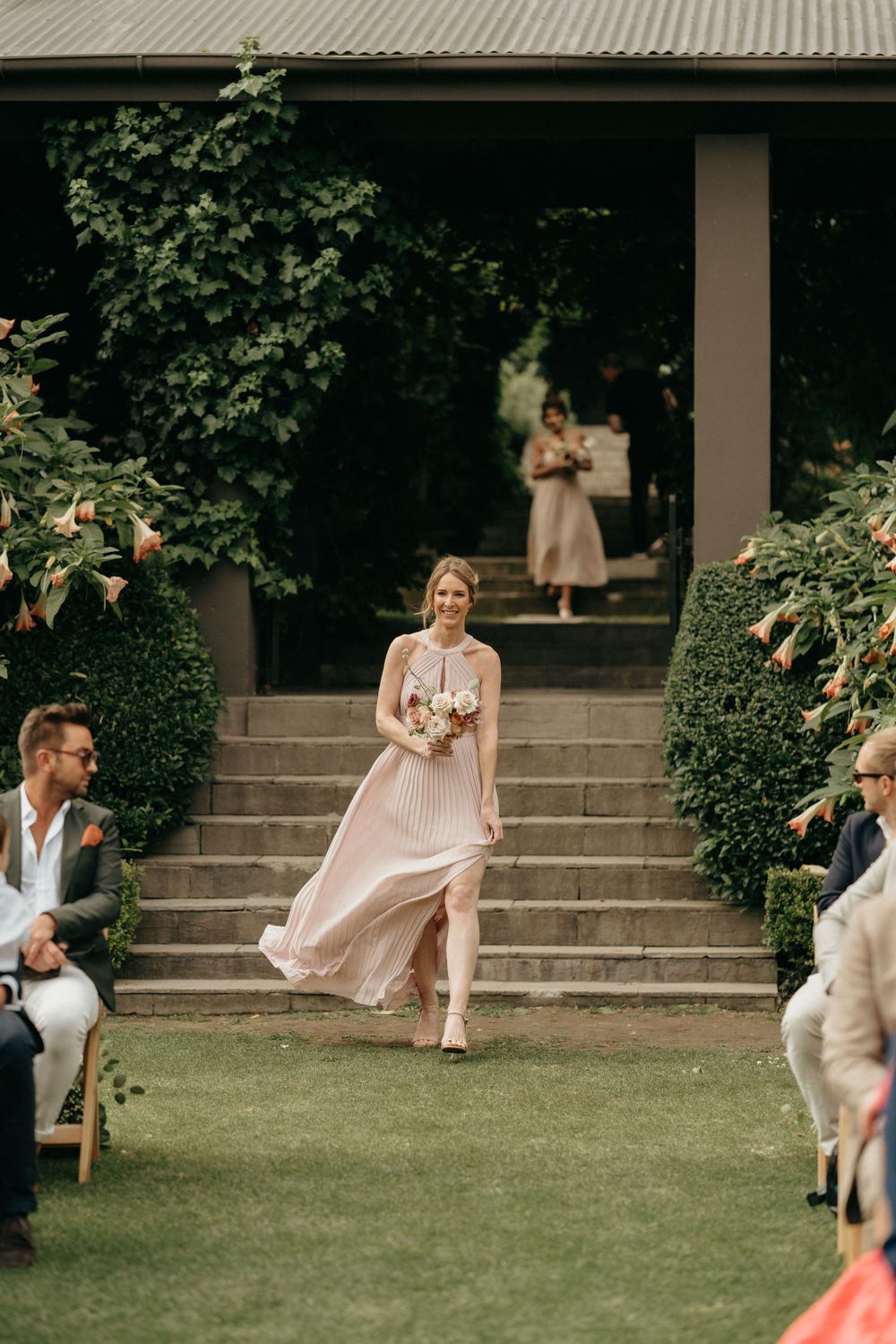 Bridesmaid in flowing blush pink gown walks down outdoor ceremony aisle with manicured garden landscaping.