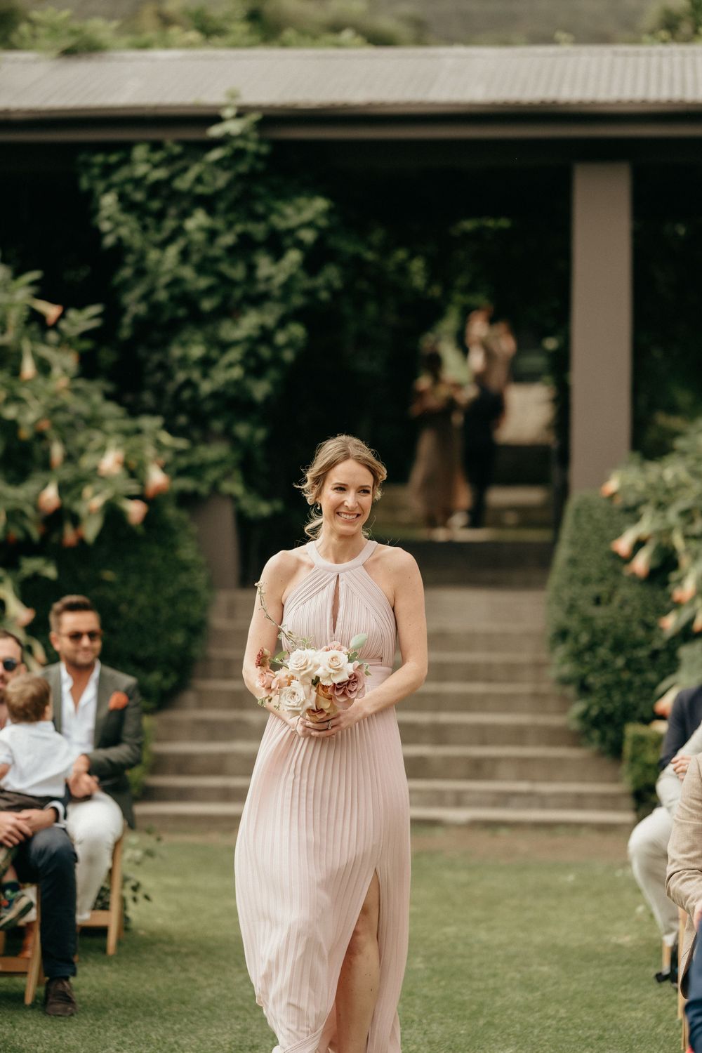 Bridesmaid wearing pleated pink halter dress carries bouquet while walking on lawn during outdoor ceremony.
