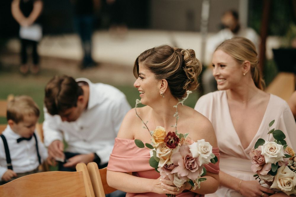 Bridesmaids in pink and blush dresses hold floral bouquets at an outdoor wedding.