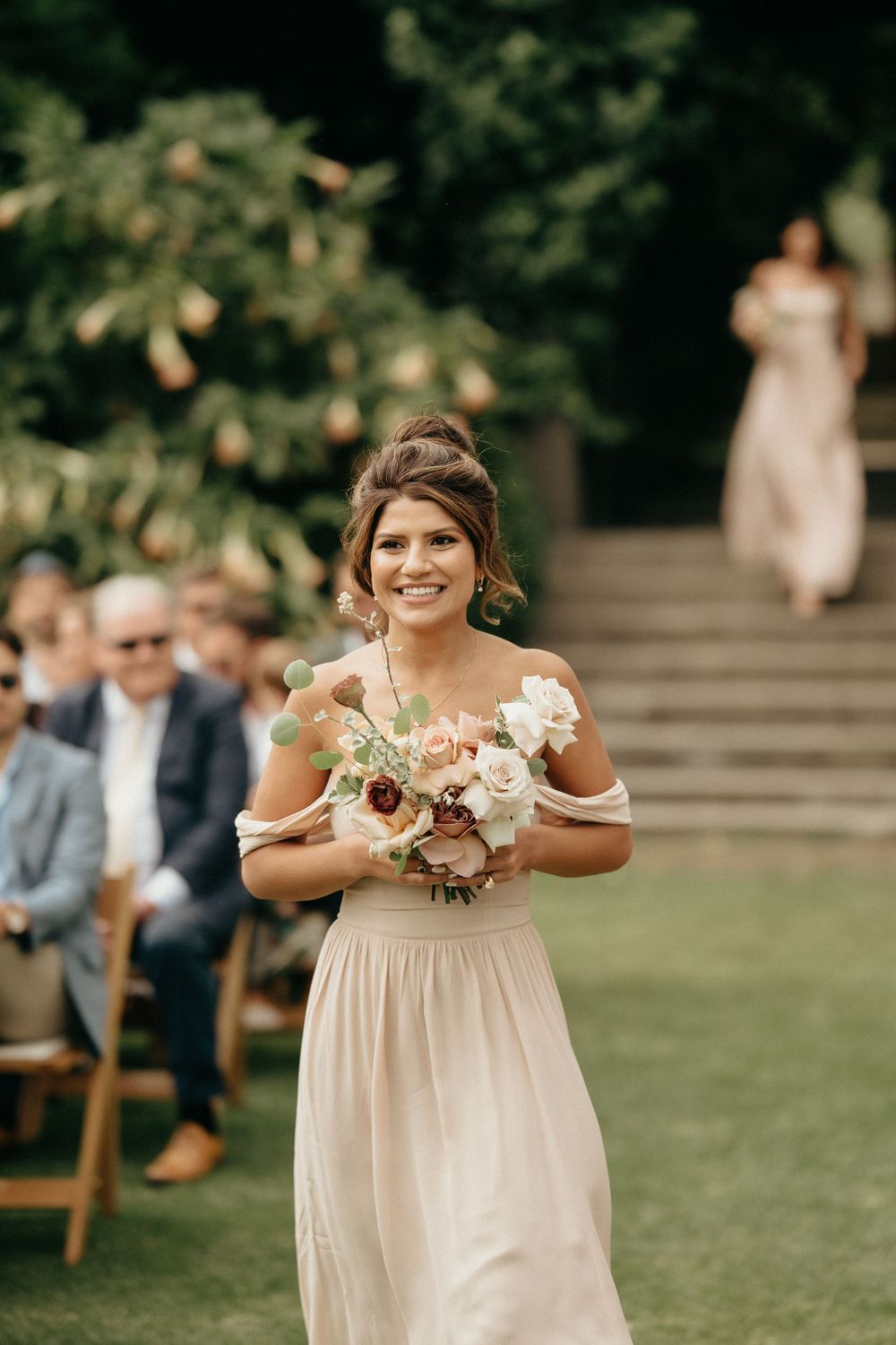 Bridesmaid in blush pink dress carrying white and burgundy floral bouquet at outdoor wedding ceremony.