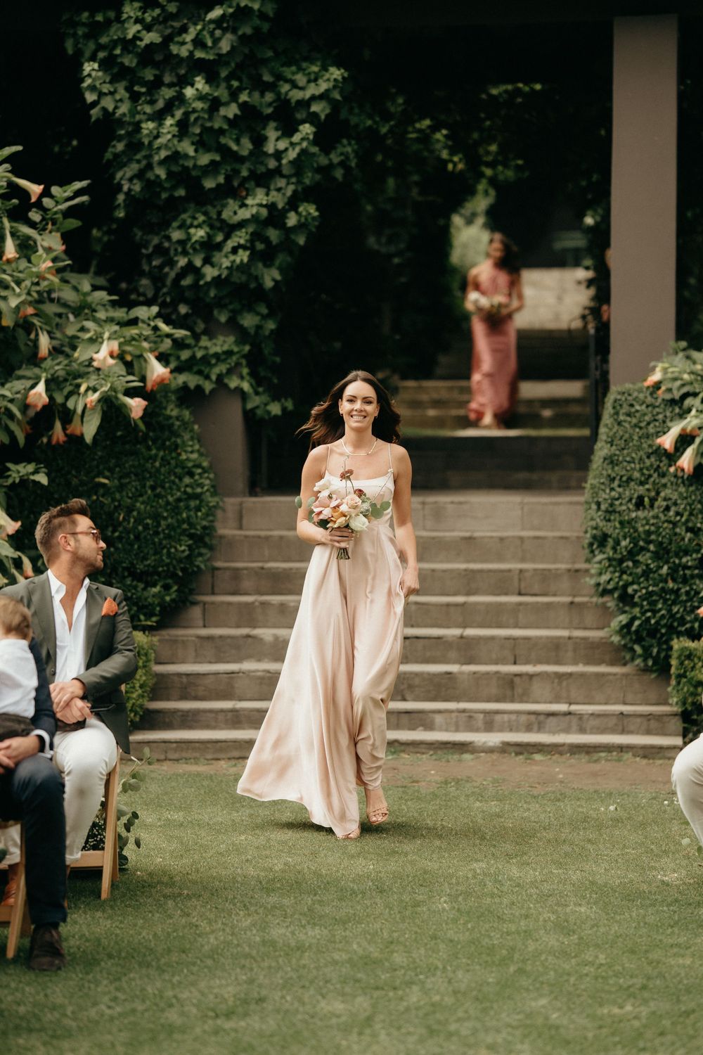 A bridesmaid in a flowing blush gown descends stone steps during an outdoor garden ceremony.