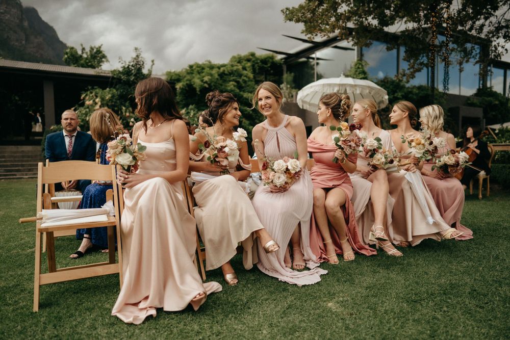 Bridesmaids wearing matching pink satin dresses sit together on chairs at an outdoor wedding ceremony.