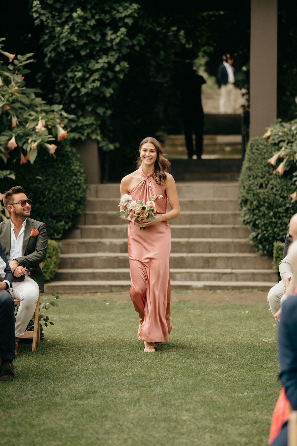 Woman in flowing pink gown walks down garden aisle holding bouquet during outdoor ceremony.