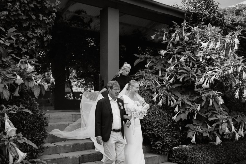 A black and white photo captures a couple exiting a columned building surrounded by lush foliage.