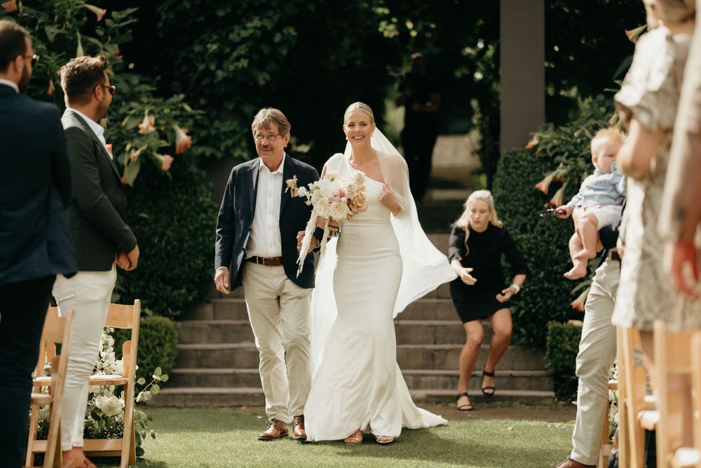 Wedding processional down an aisle lined with wooden folding chairs in a garden setting.