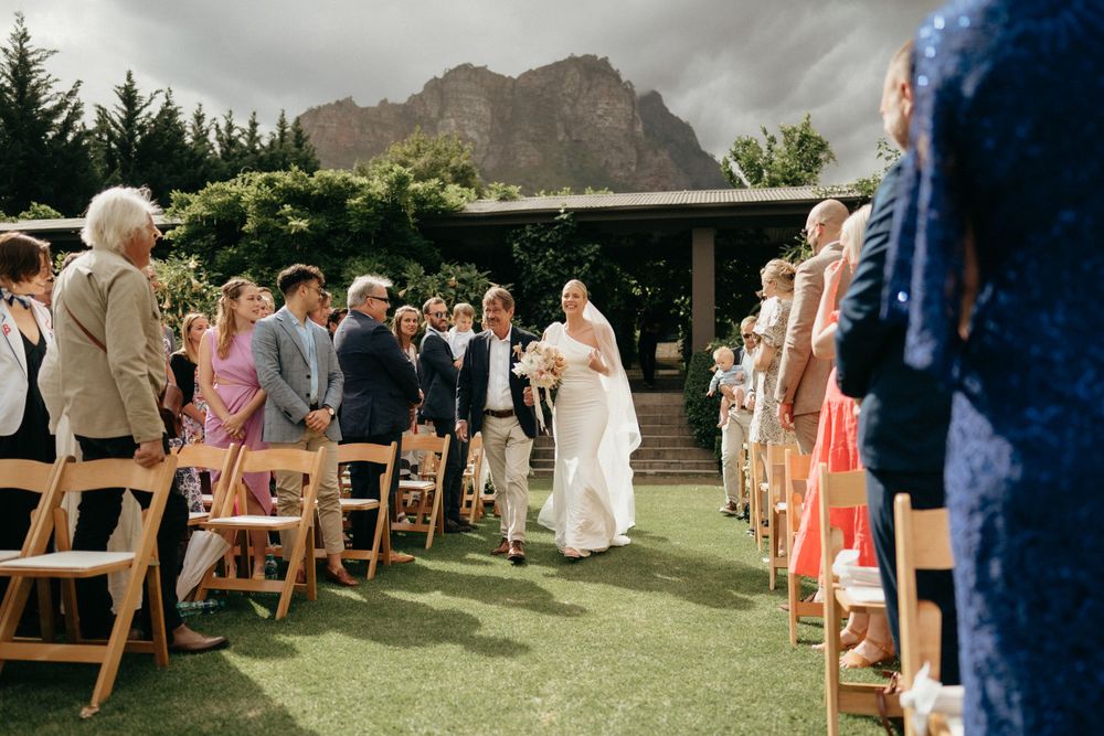 Outdoor wedding ceremony with guests seated on folding chairs against a mountainous landscape.