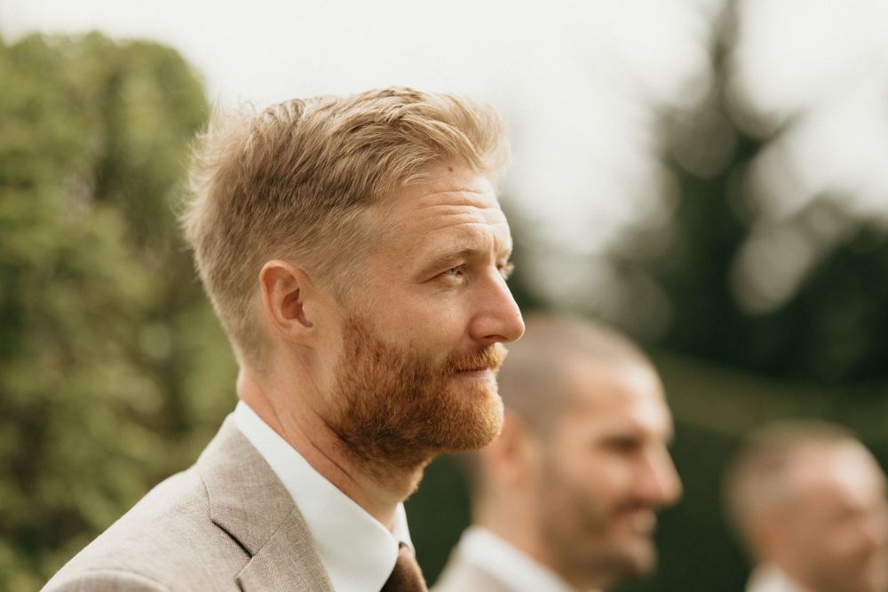 A wedding attendee in a light brown suit looks on during an outdoor ceremony.
