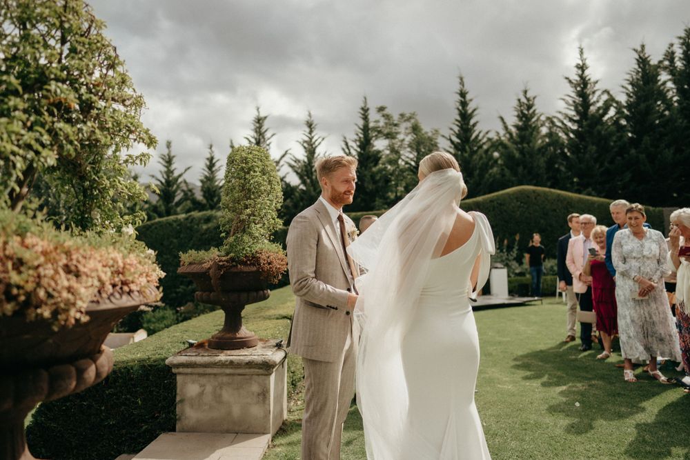 An outdoor garden wedding ceremony with guests watching a couple exchange vows among evergreen trees and manicured landscaping.