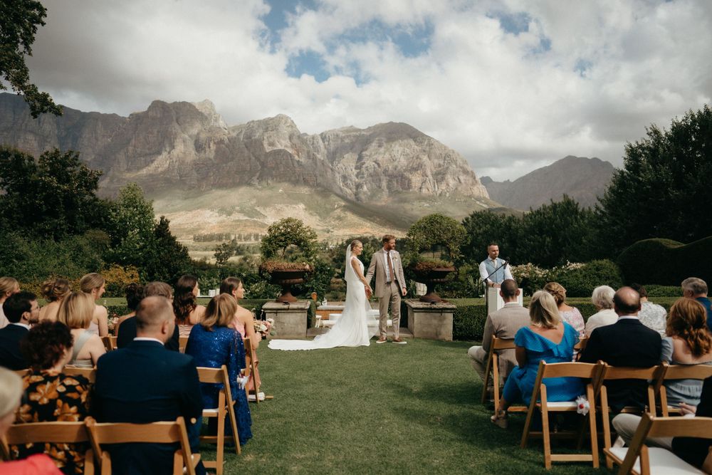 Wedding ceremony setup with wooden chairs on lawn against dramatic mountain backdrop under cloudy sky.