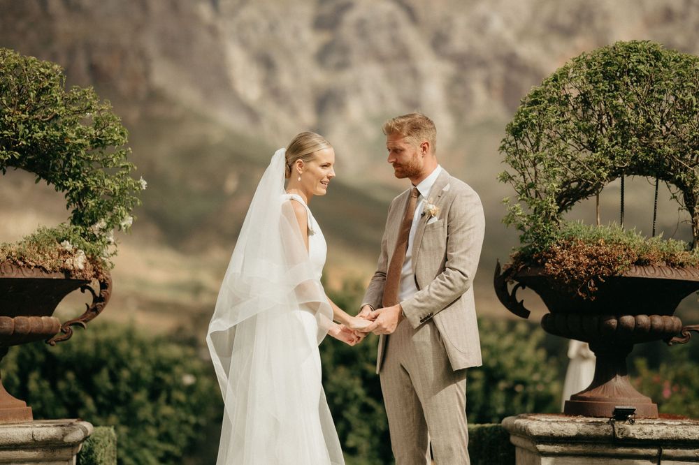 A wedding couple shares an intimate moment in front of decorative garden urns with dramatic mountain backdrop.