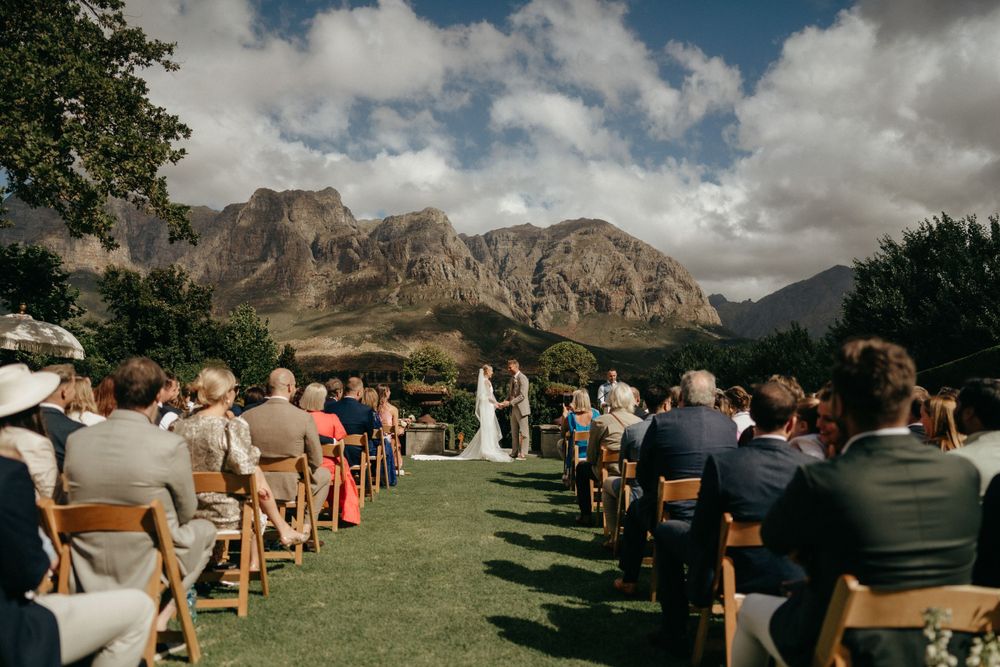 An outdoor wedding ceremony set against dramatic mountain peaks with guests seated on a grassy lawn.