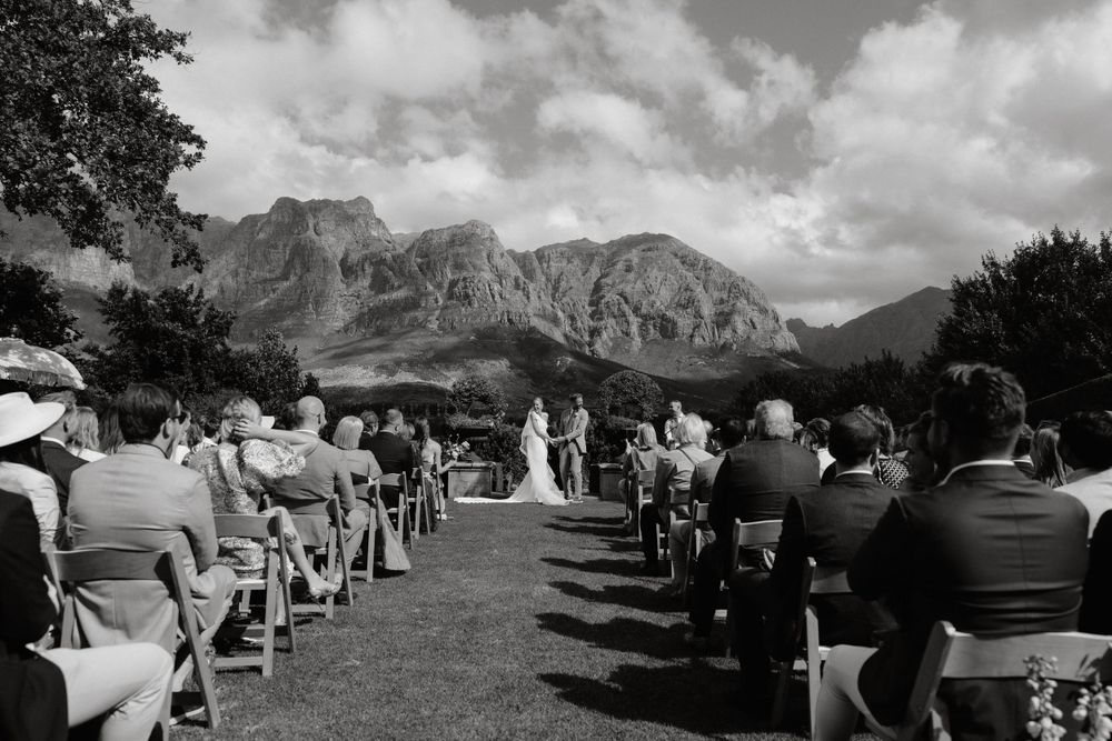 An outdoor wedding ceremony with mountain views in the background and guests seated in rows of white chairs.