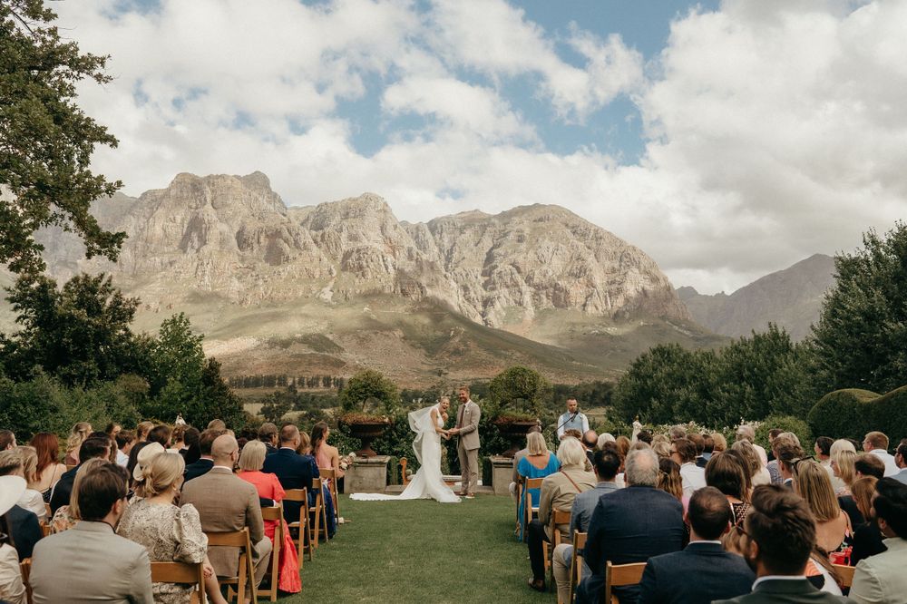 Outdoor mountain wedding ceremony with guests seated on white chairs facing majestic rocky peaks under blue skies.