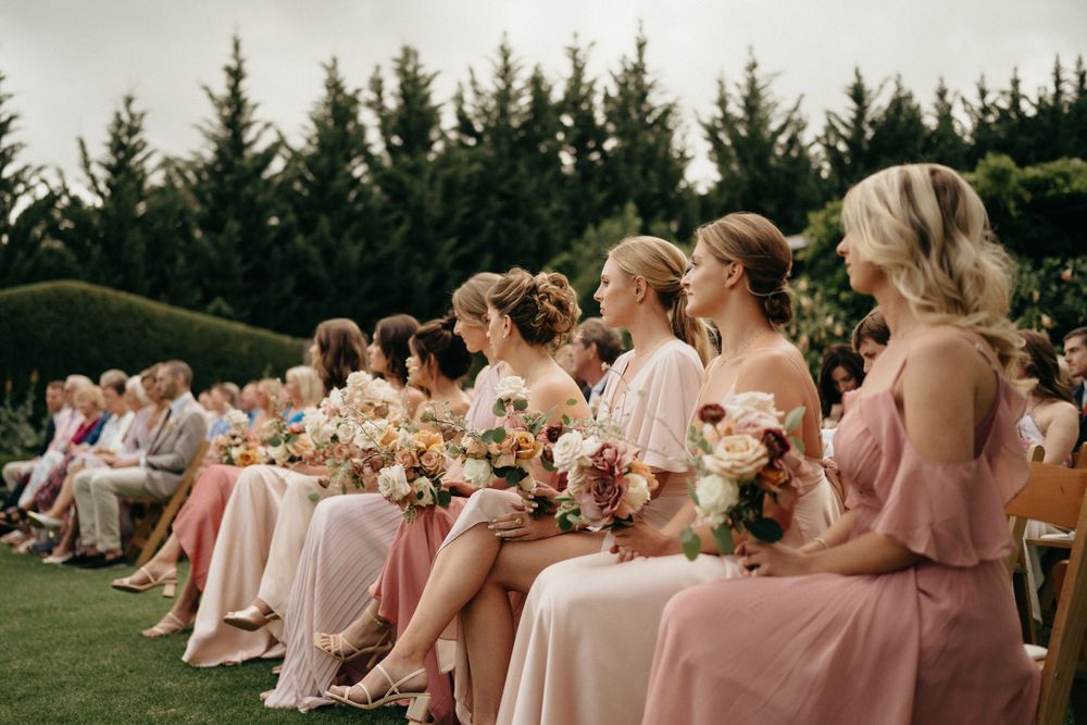 Bridesmaids wearing pink dresses sit on a row holding floral bouquets during an outdoor ceremony.