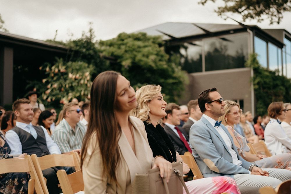 Wedding guests seated in rows of wooden chairs at an outdoor ceremony with modern architecture.