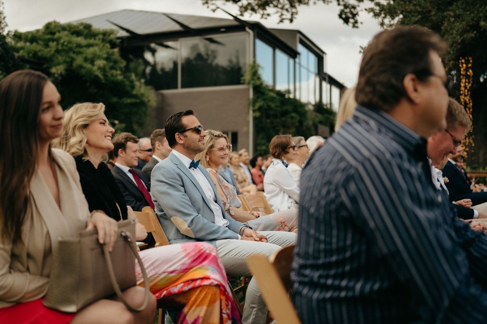 Guests seated on wooden chairs at outdoor wedding with modern building in background.
