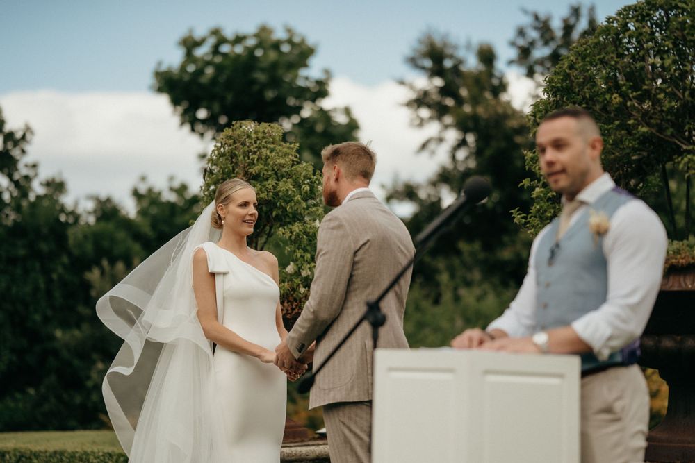 A wedding ceremony takes place outdoors with attendees gathered under trees on a sunny day.