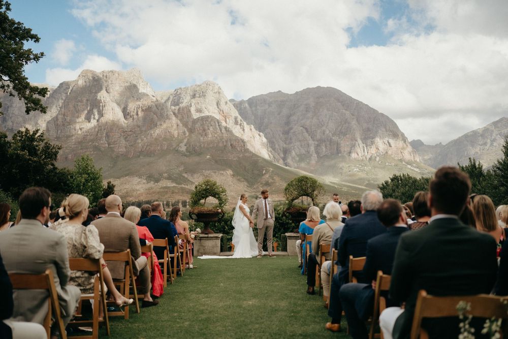 A wedding ceremony takes place outdoors with dramatic mountain peaks in the background and guests seated on wooden chairs.