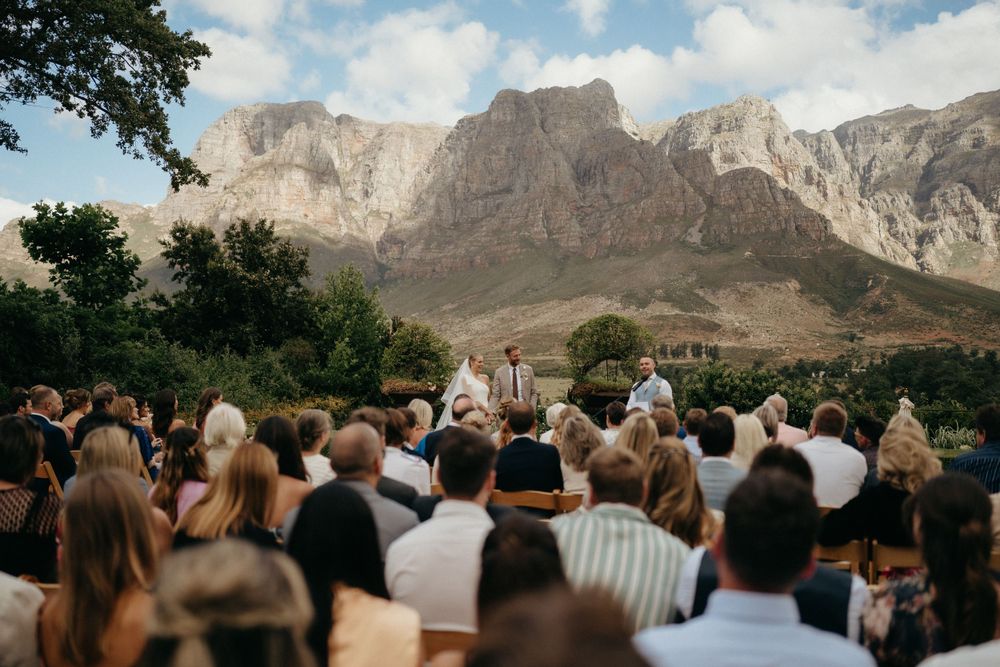 Wedding ceremony amid stunning mountain scenery with guests watching from outdoor seating area.