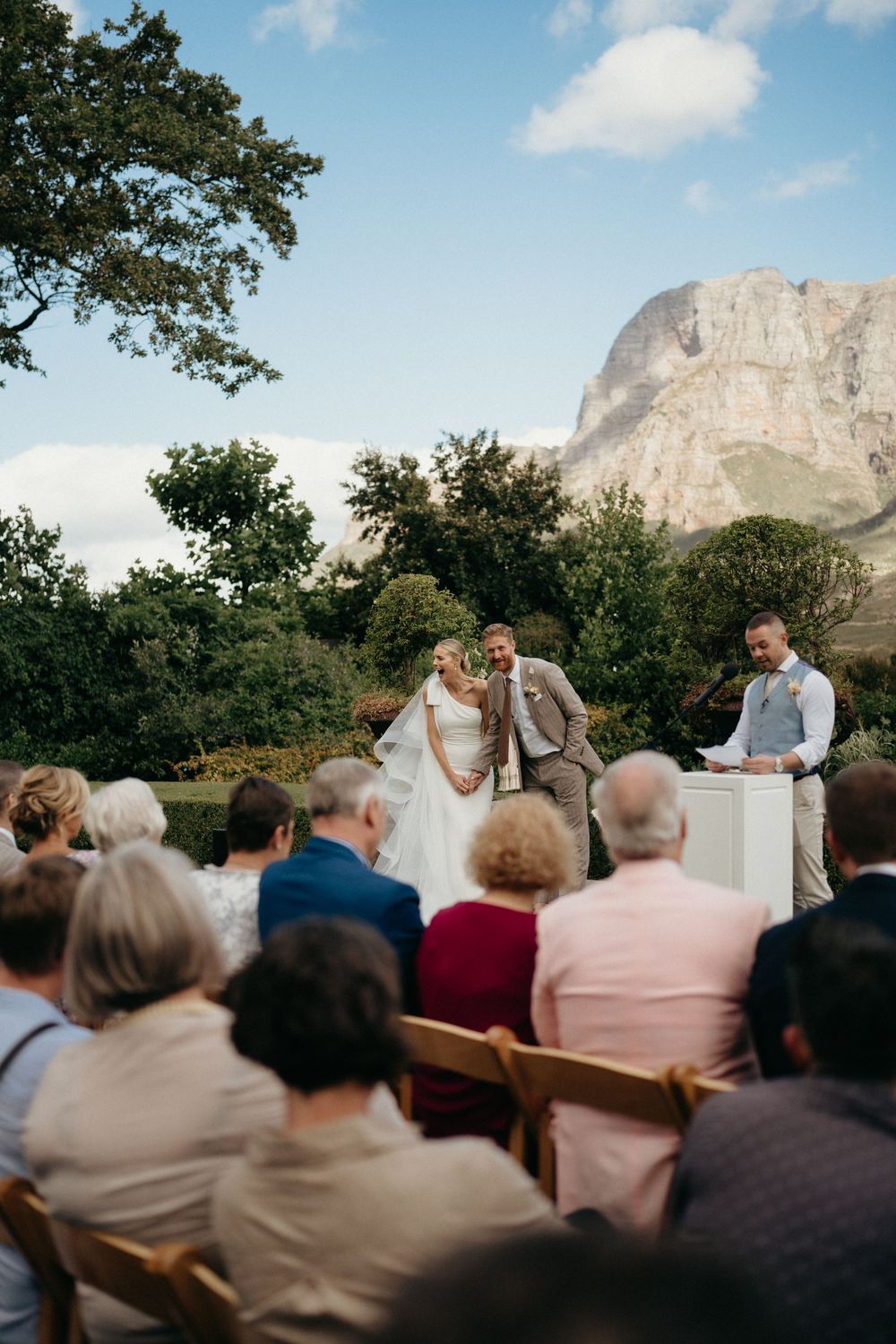 An outdoor wedding ceremony takes place against a dramatic mountain backdrop with guests seated on wooden chairs.