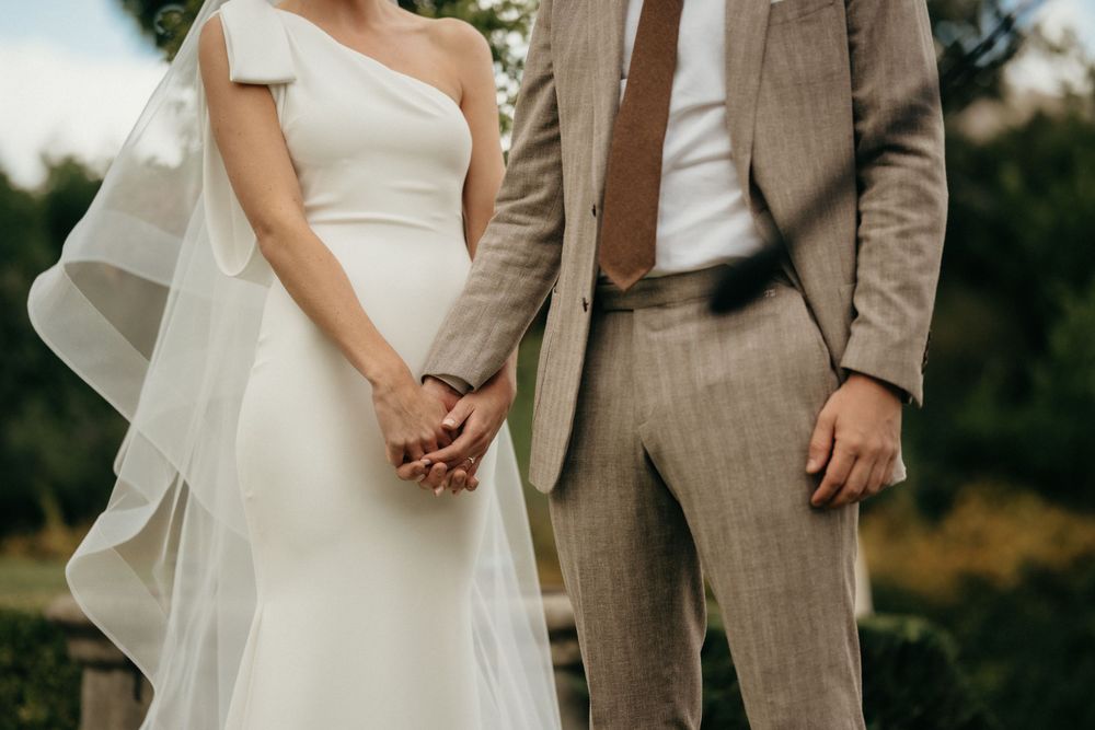 A bride in white gown and groom in tan suit hold hands during their wedding ceremony.
