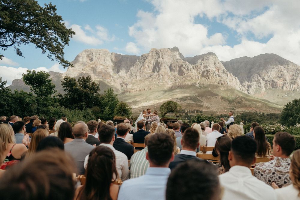 An outdoor wedding ceremony with guests gathered against a backdrop of dramatic mountain peaks and blue skies.