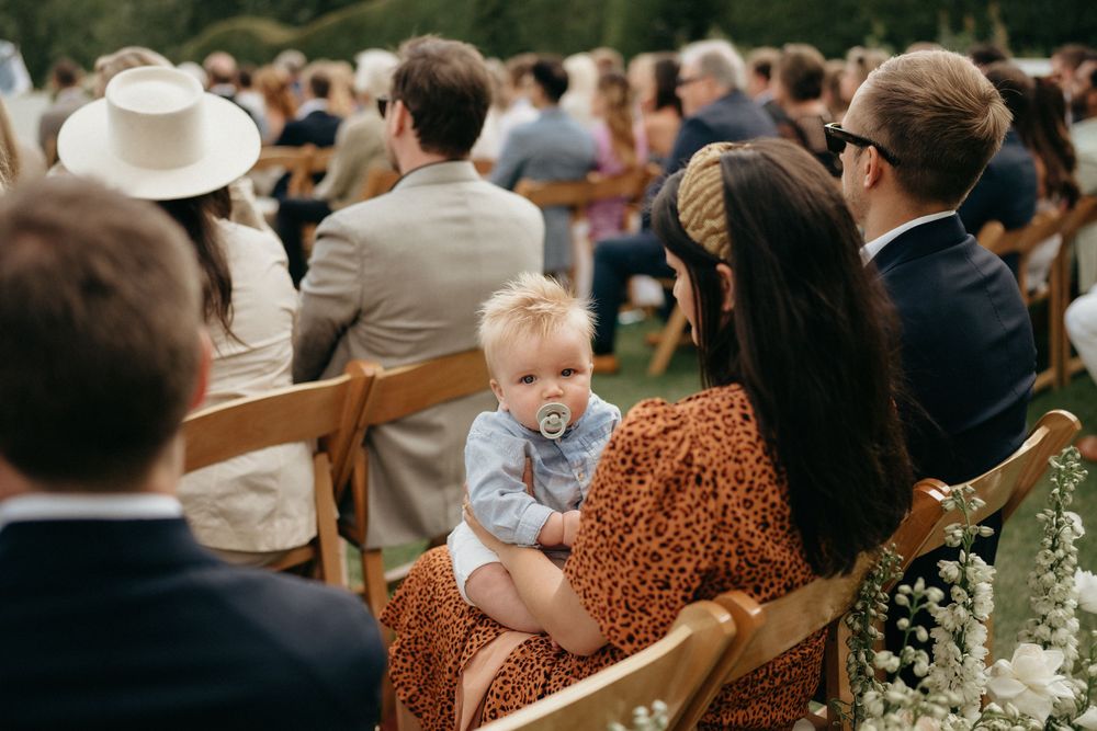 Happy guests seated in wooden chairs at an outdoor wedding ceremony with greenery in the background.