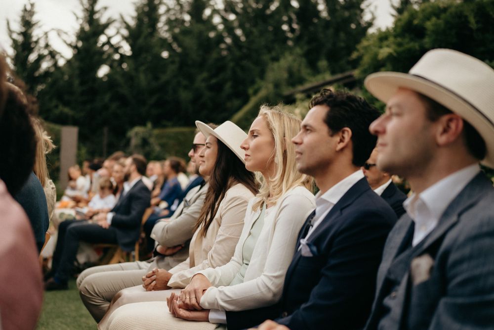 Wedding guests in summer attire seated on wooden chairs during an outdoor ceremony.